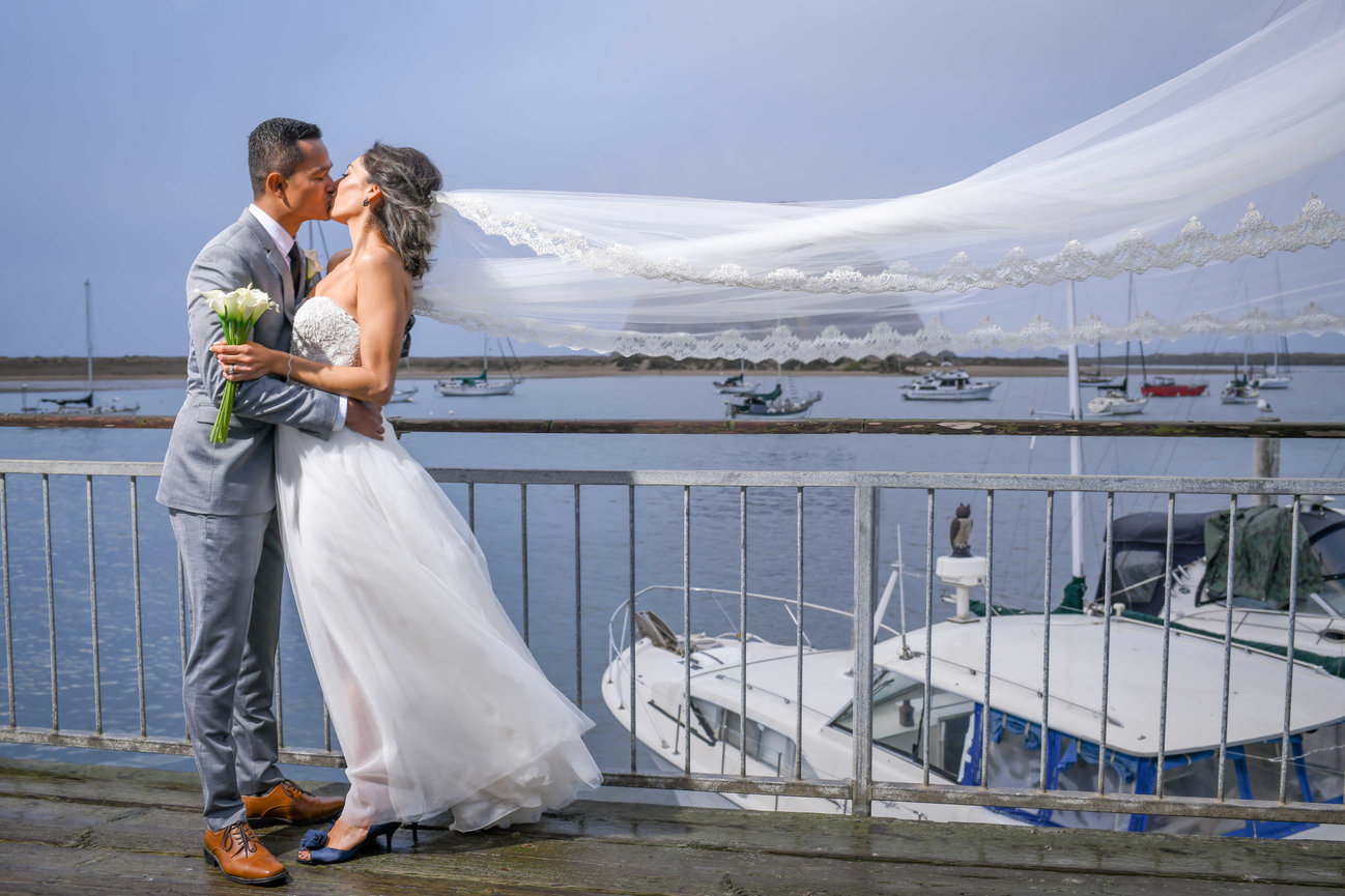 A kiss on the boards with the veil thrown sideways by the wind off the water.